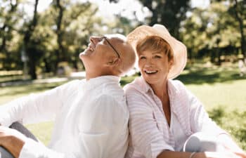 Trendy short haired lady in light hat and striped blouse smiling and sitting on grass with old man in glasses and white shirt outdoor.